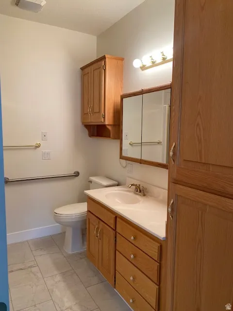 Light-toned wood-finish cabinetry, white countertop with integrated sink, rectangular wall-mounted mirror, light-toned tile flooring, and wall-mounted grab bars