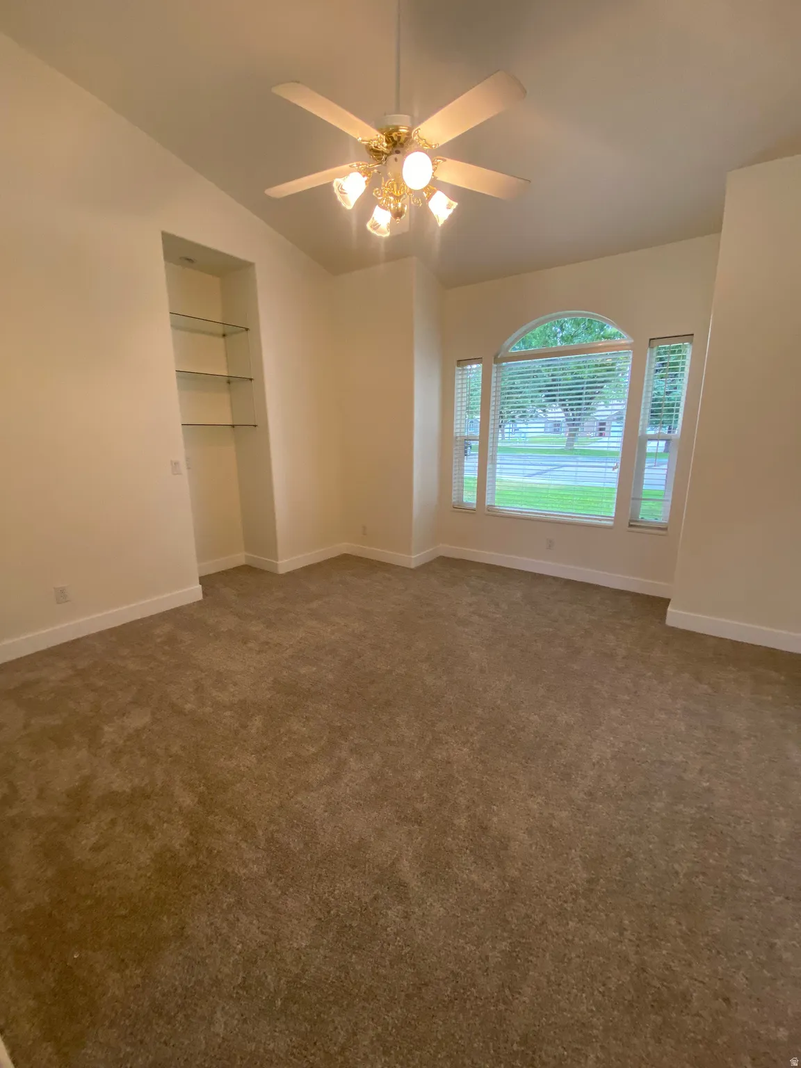 Arched window with blinds, ceiling fan with integrated lighting, built-in recessed shelving with glass shelves, and vaulted ceilings