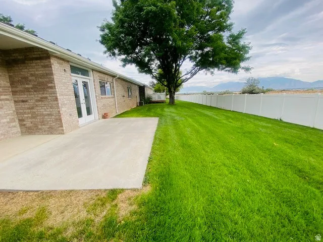 Expansive green lawn with mature tree, concrete patio, brick exterior, white privacy fence, and double French doors
