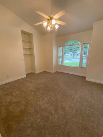 Arched window with blinds, ceiling fan with integrated lighting, built-in recessed shelving with glass shelves, and vaulted ceilings