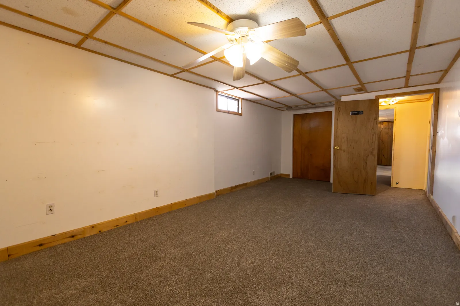 Carpeted room featuring a drop ceiling with wood-finish grid, a ceiling fan with integrated lighting, wood-finish baseboards, and a wood-finish door