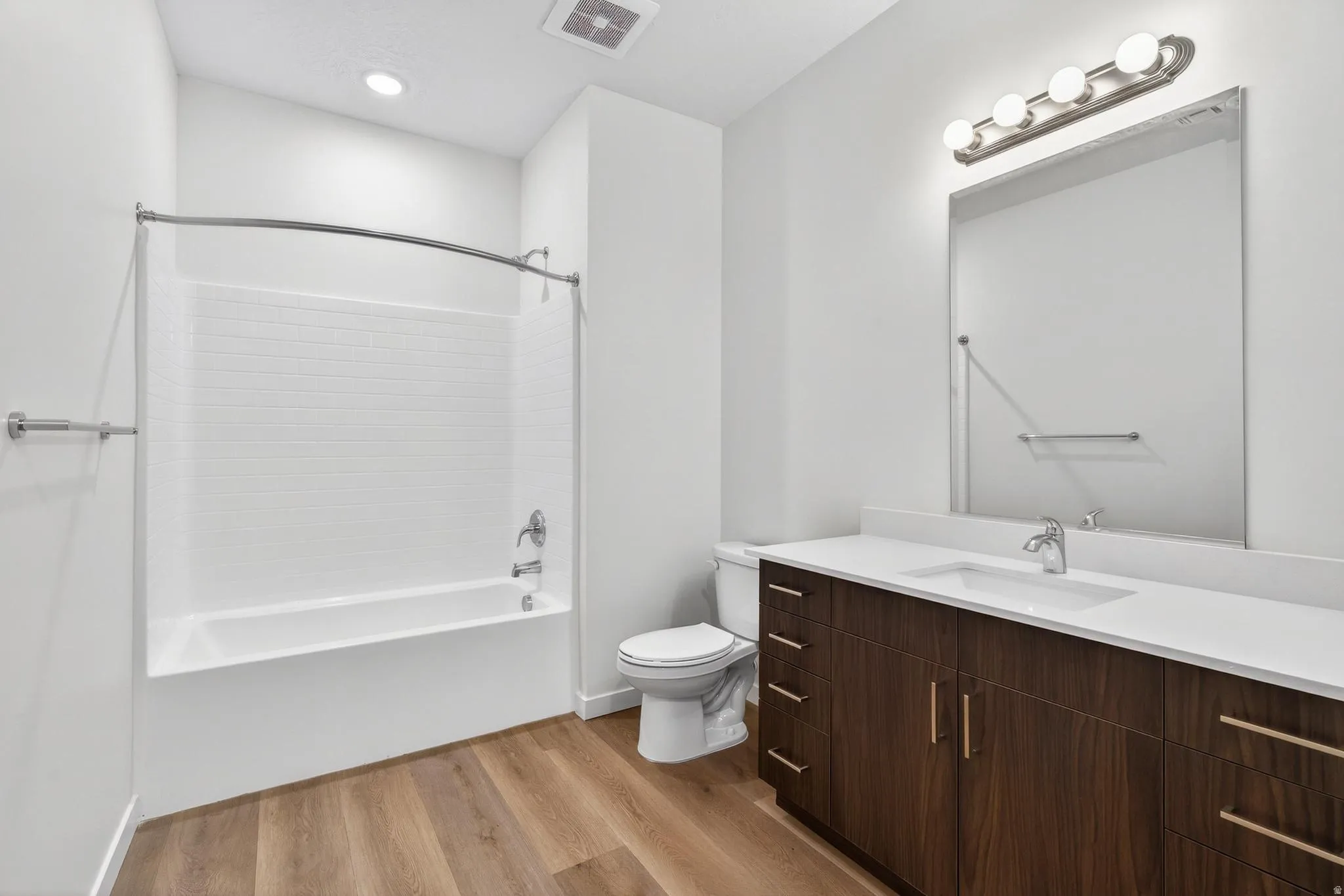 Bathroom featuring wood-finish flooring, a white subway tile tub surround, an elongated toilet, a dark wood-finish vanity with a white countertop and undermount sink, and a large mirror with overhead lighting