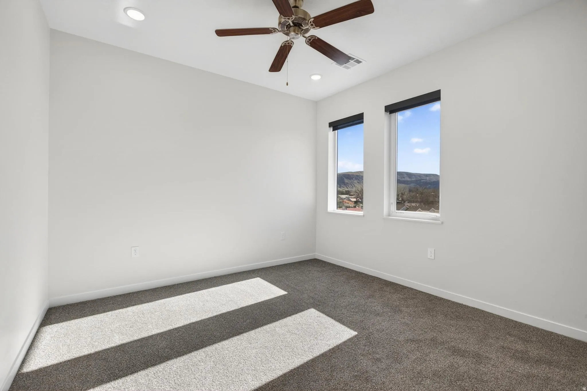 Bright room featuring two windows with roller shades, a ceiling fan, recessed lighting, and grey carpet flooring