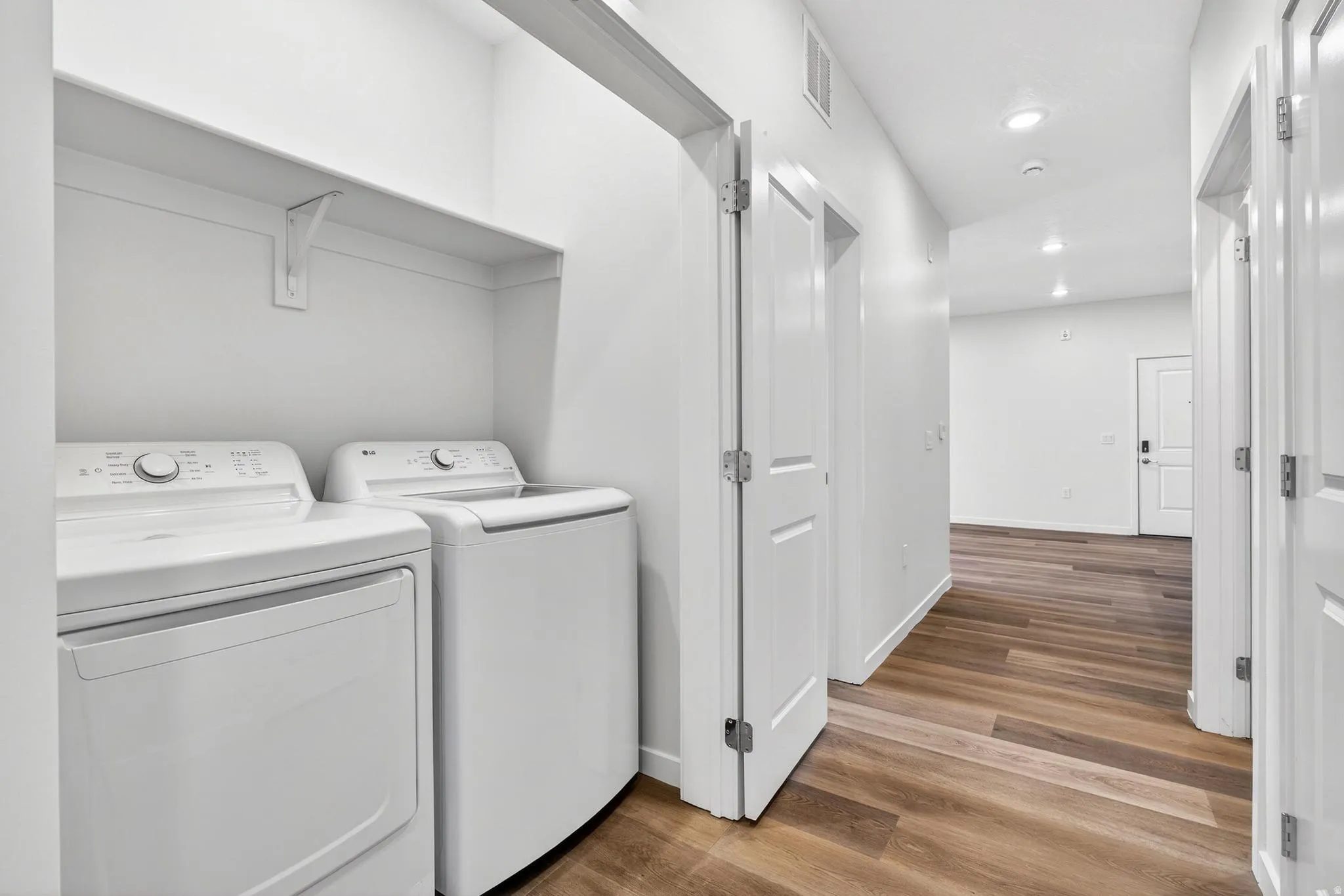 Dedicated laundry area featuring a built-in shelf, adjacent to a hallway with wood-finish flooring, recessed lighting, and multiple white doors