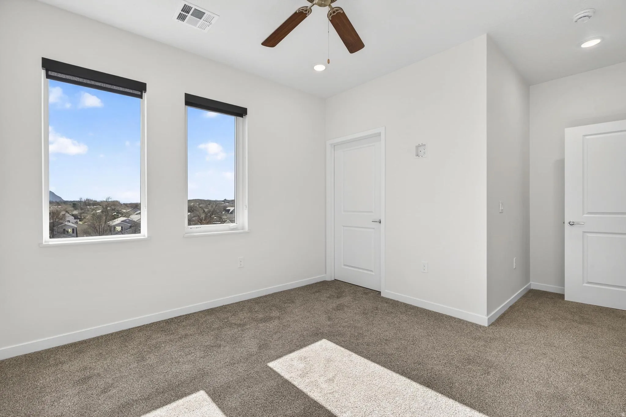 Room featuring two windows with roller shades, white walls, and neutral carpet flooring