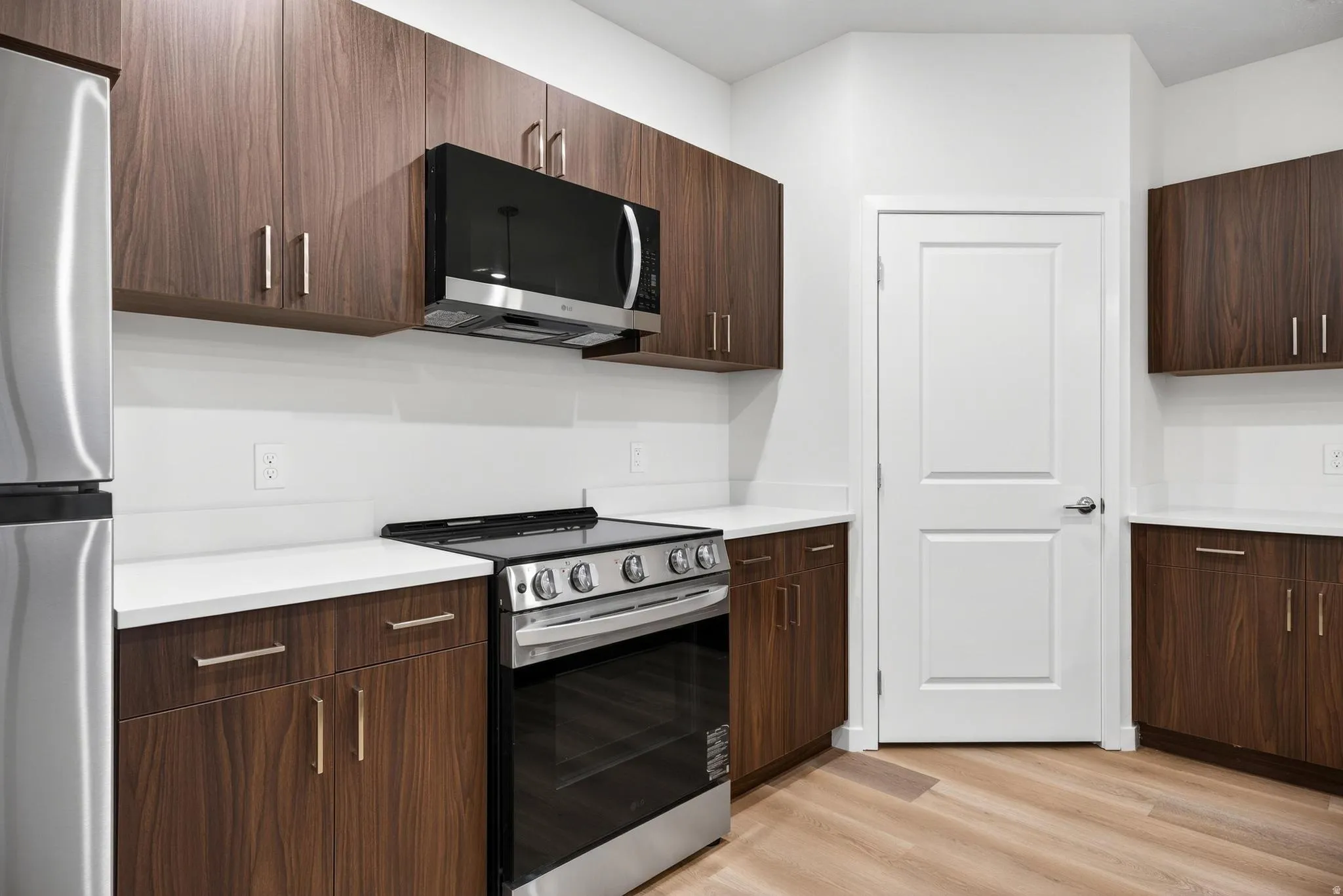 Modern kitchen featuring dark wood-finish cabinetry with brushed nickel hardware, white countertops, and stainless steel appliances including a refrigerator, range, and microwave