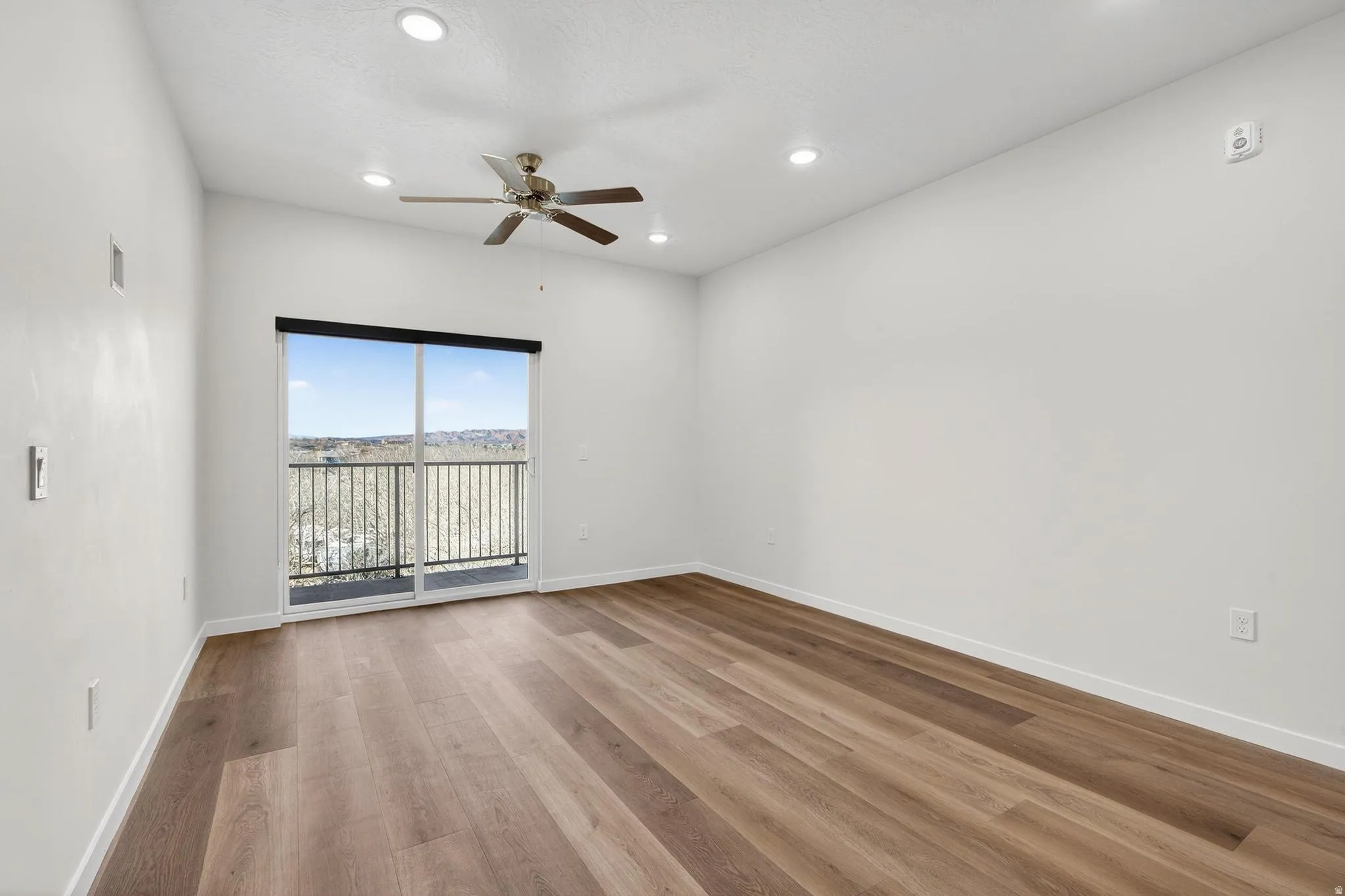 Bright interior space featuring wood-finish flooring, a sliding glass door to a balcony with metal railings, a ceiling fan with integrated lighting, and recessed ceiling lights
