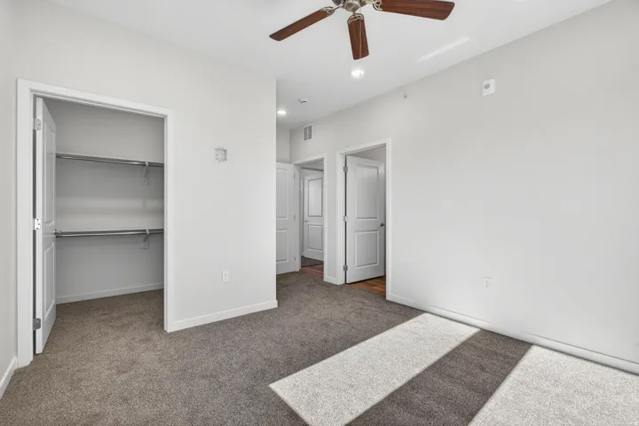 Carpeted interior space featuring a ceiling fan with wood-finish blades, a white paneled door, and a closet with shelving and hanging rods