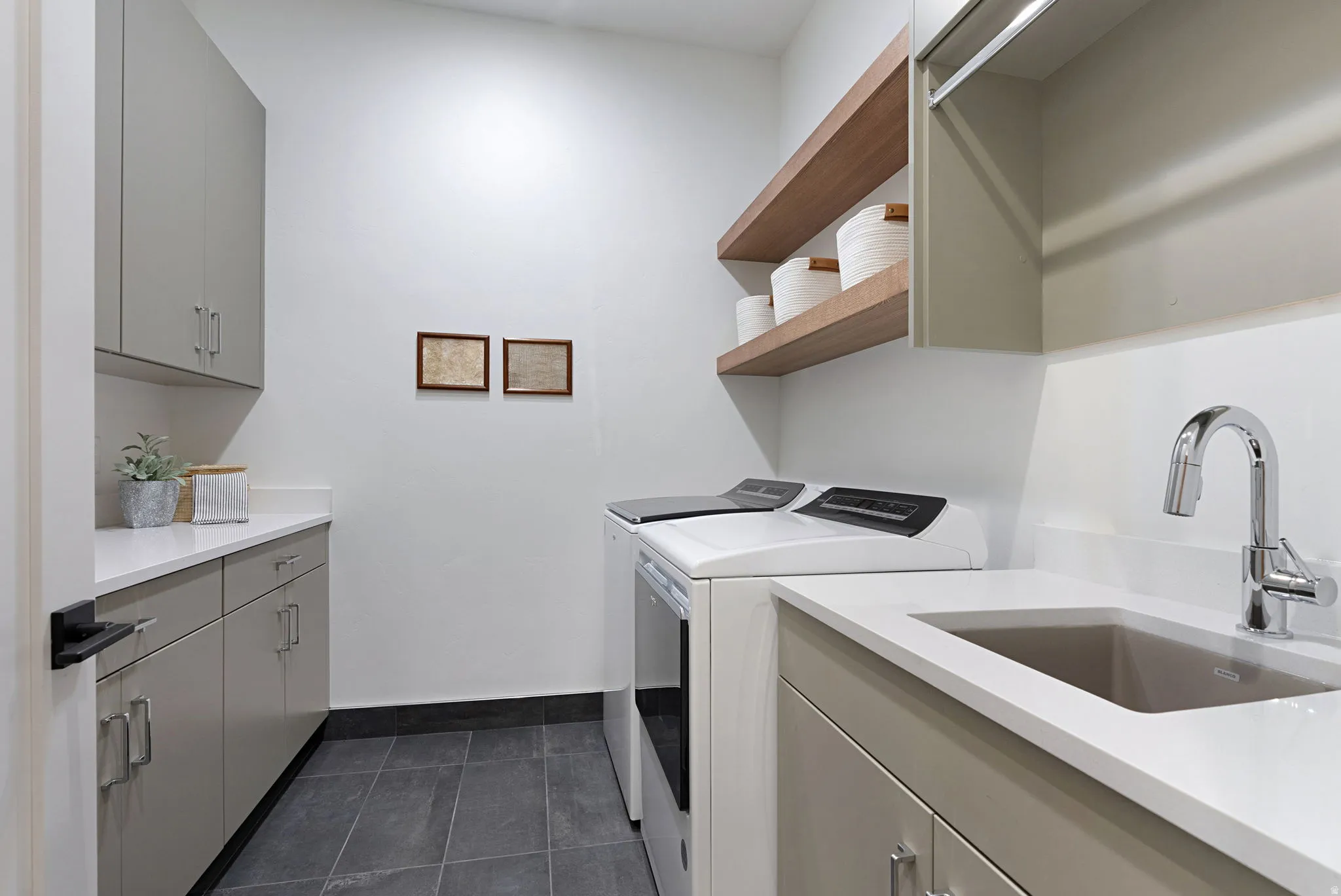 Dedicated utility space featuring a built-in sink with chrome faucet, light grey cabinetry, white countertops, dark tile flooring, and open wood shelving