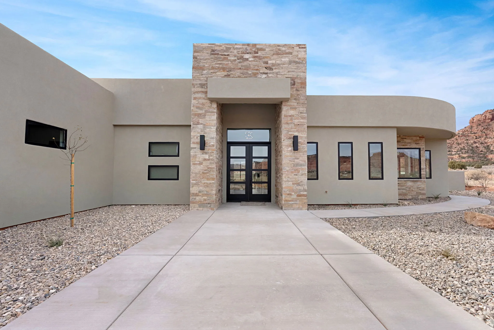 Modern stucco exterior featuring a stone facade entry, a glass-paneled entry door, black-framed windows, and a curved architectural element