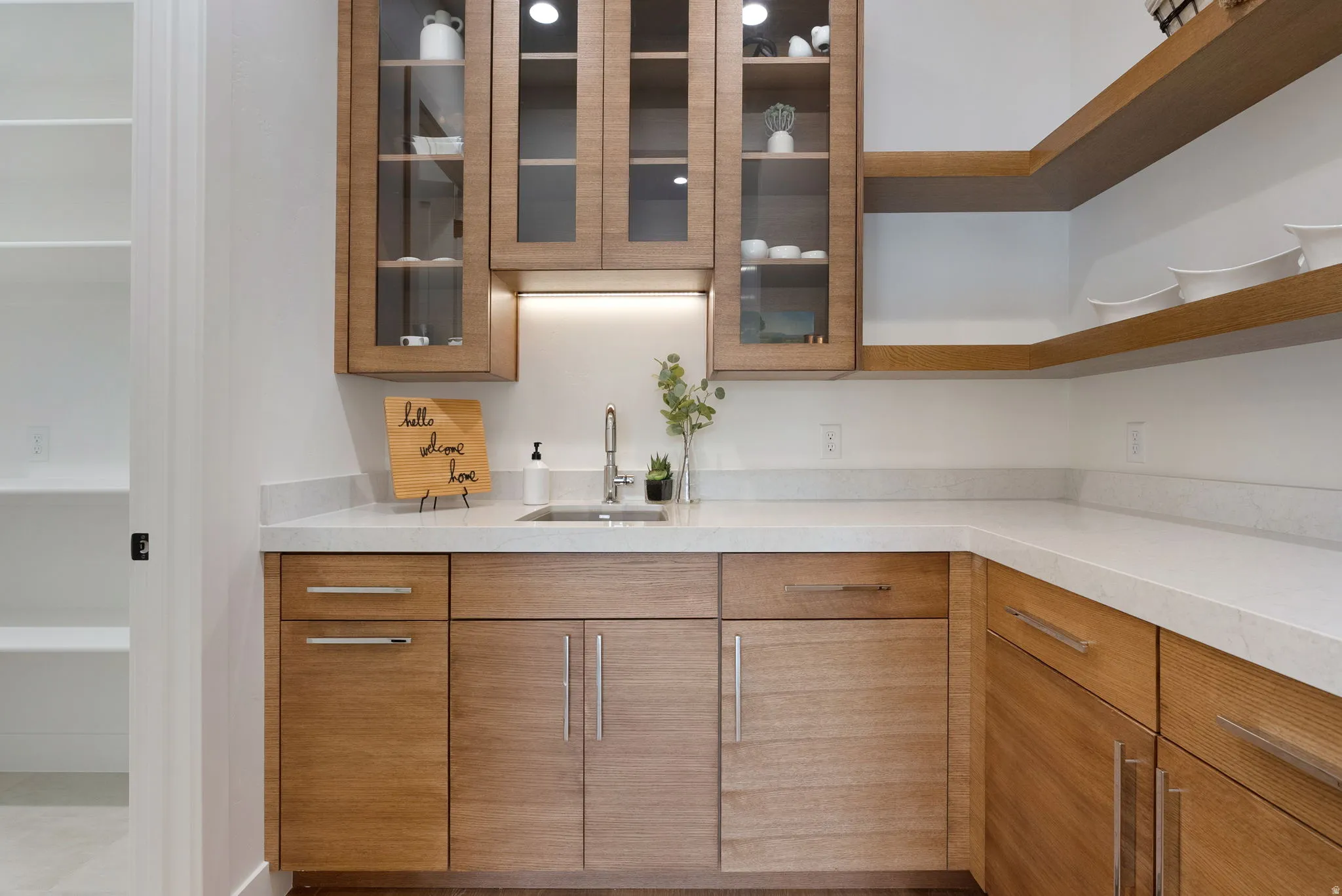 Kitchenette featuring wood cabinetry, white stone countertops, a stainless steel sink with a gooseneck faucet, glass-front upper cabinets, and open wood shelving