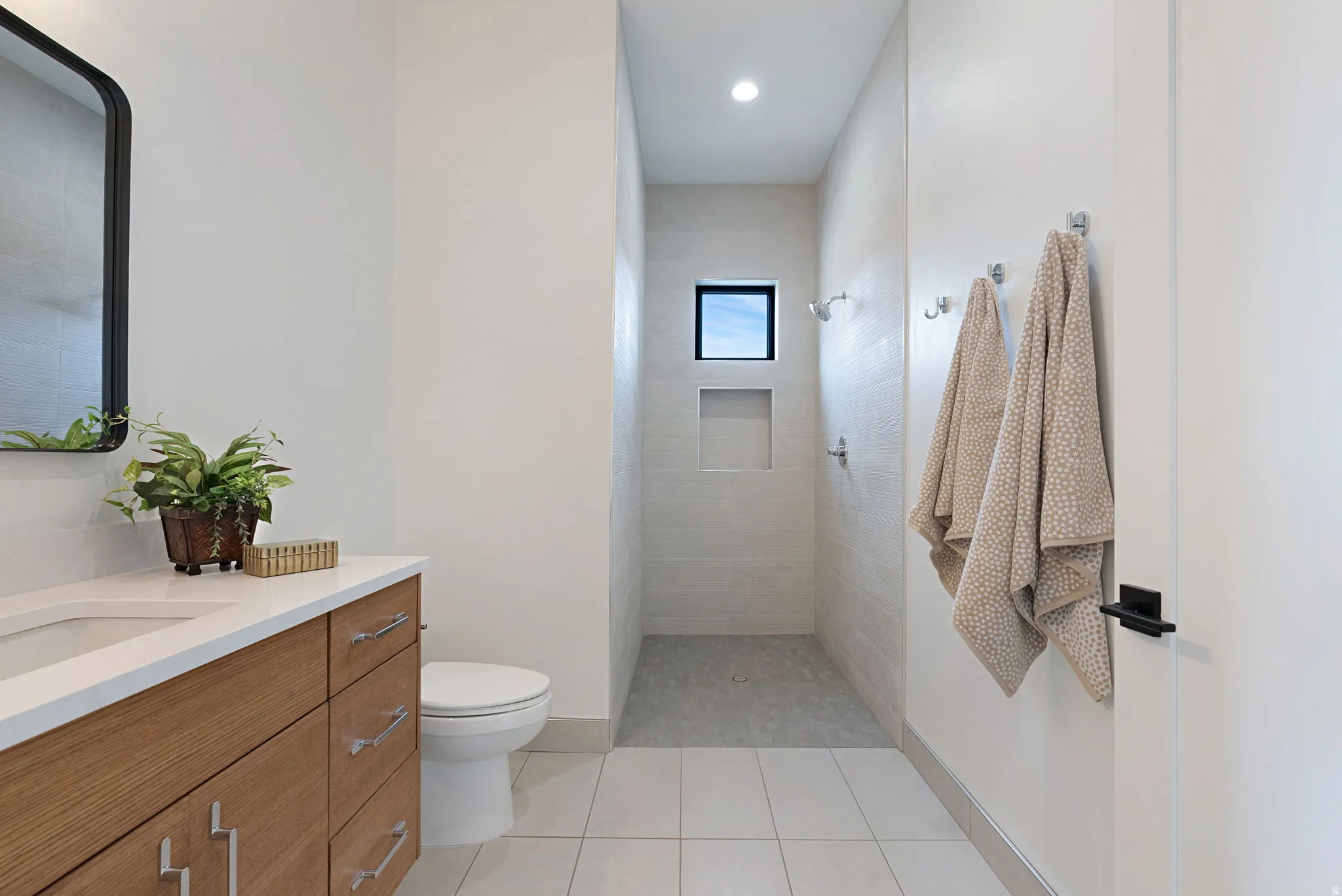 Modern bathroom featuring a wood-finish vanity with a white countertop and undermount sink, a sleek black-framed mirror, and a walk-in shower with light-colored tile walls, a built-in niche, and a small window