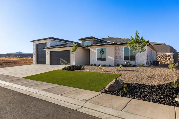 Contemporary stucco and stone-clad residence featuring a two-car garage, expansive driveway, and low-maintenance desert landscaping with artificial turf accents