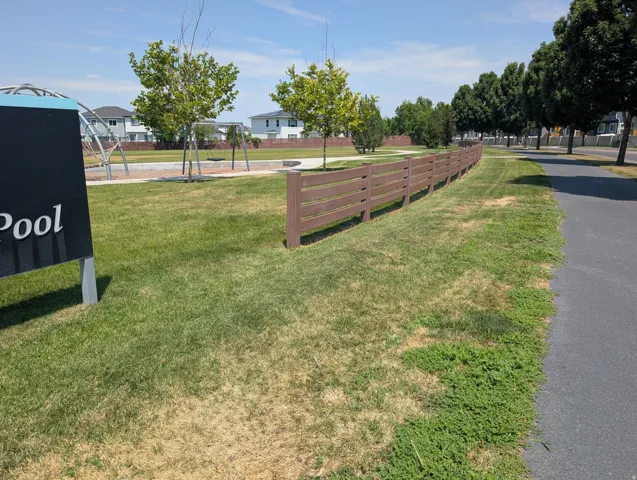 Community green space featuring a paved pathway, mature trees, and a slatted brown fence
