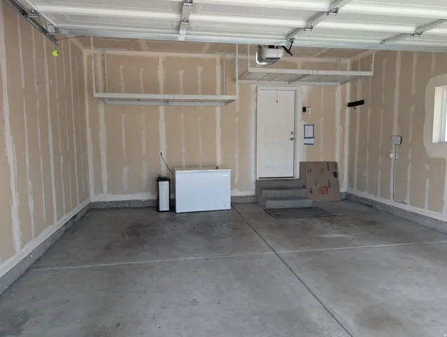 Spacious garage featuring a concrete floor, exposed drywall with visible seams, two overhead storage racks, a white access door, and a built-in window