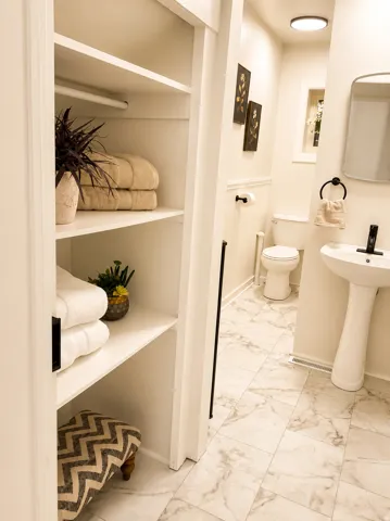 Built-in shelving, pedestal sink, wall-mounted mirror, marble-look tile flooring, and flush-mount ceiling light