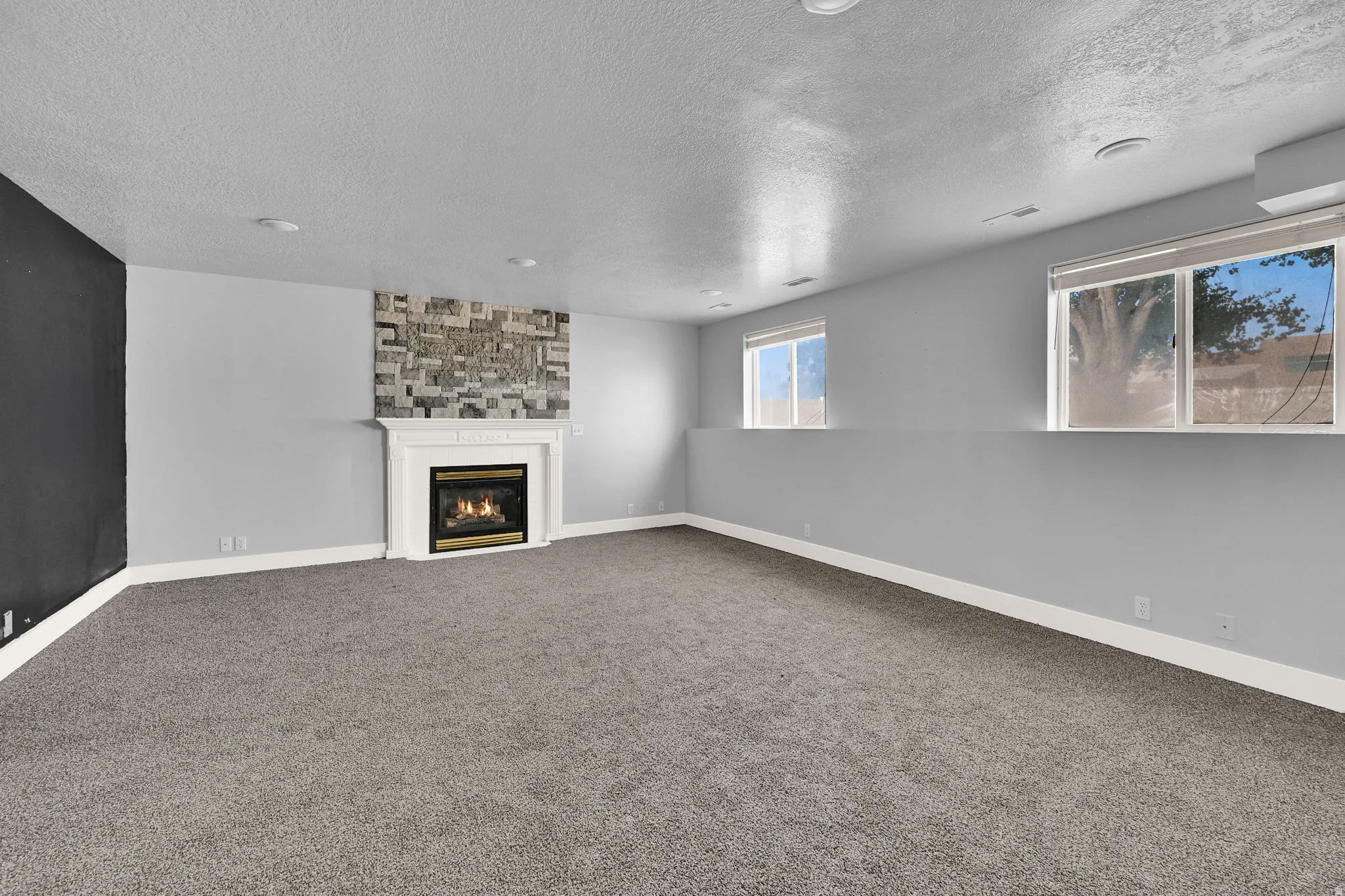 Spacious room featuring a white-mantled fireplace with a decorative stone surround, recessed lighting, and soft gray carpeting