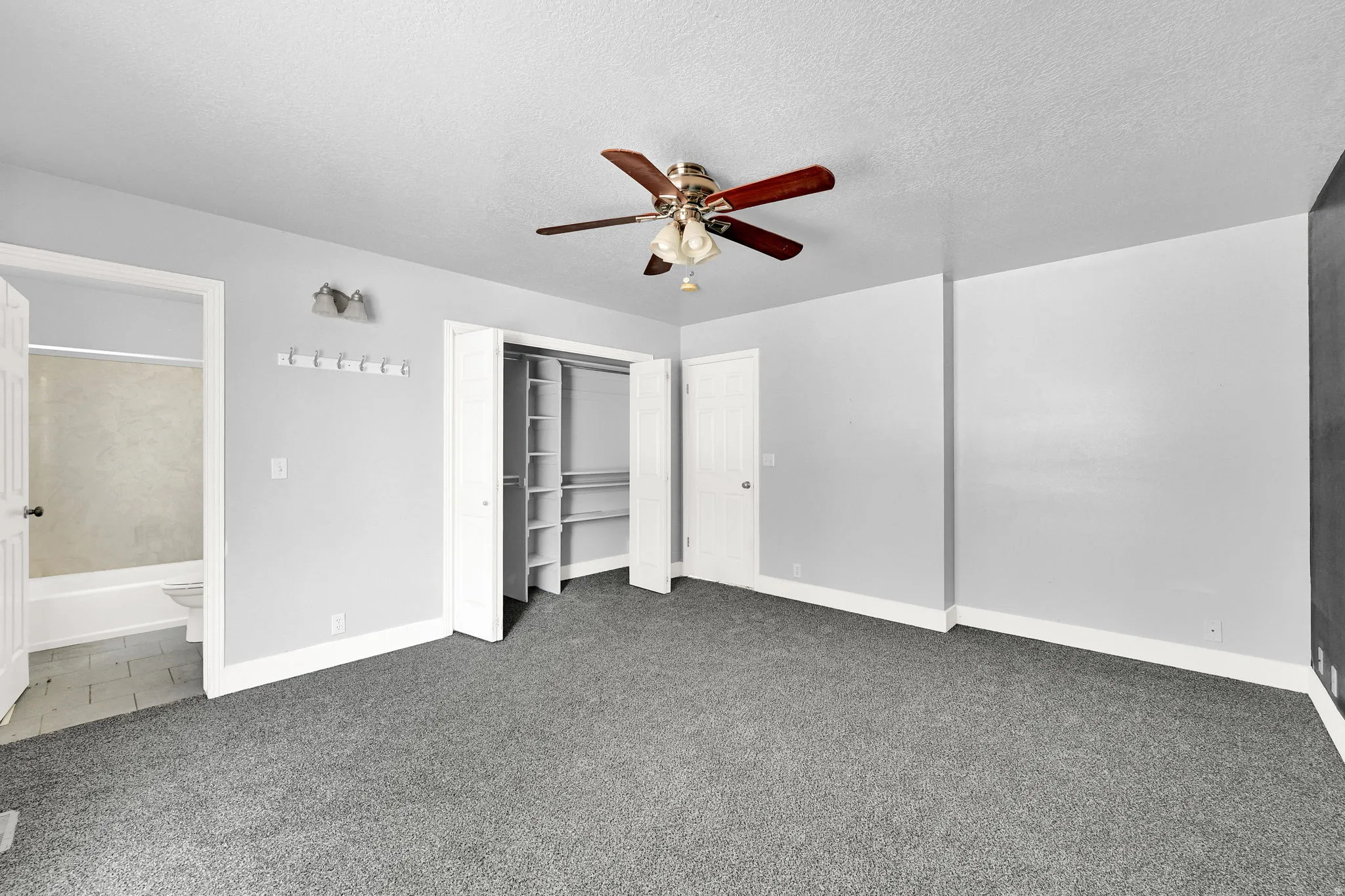 Spacious room featuring gray carpeting, light gray walls, a ceiling fan with light fixture, and a built-in closet with shelving