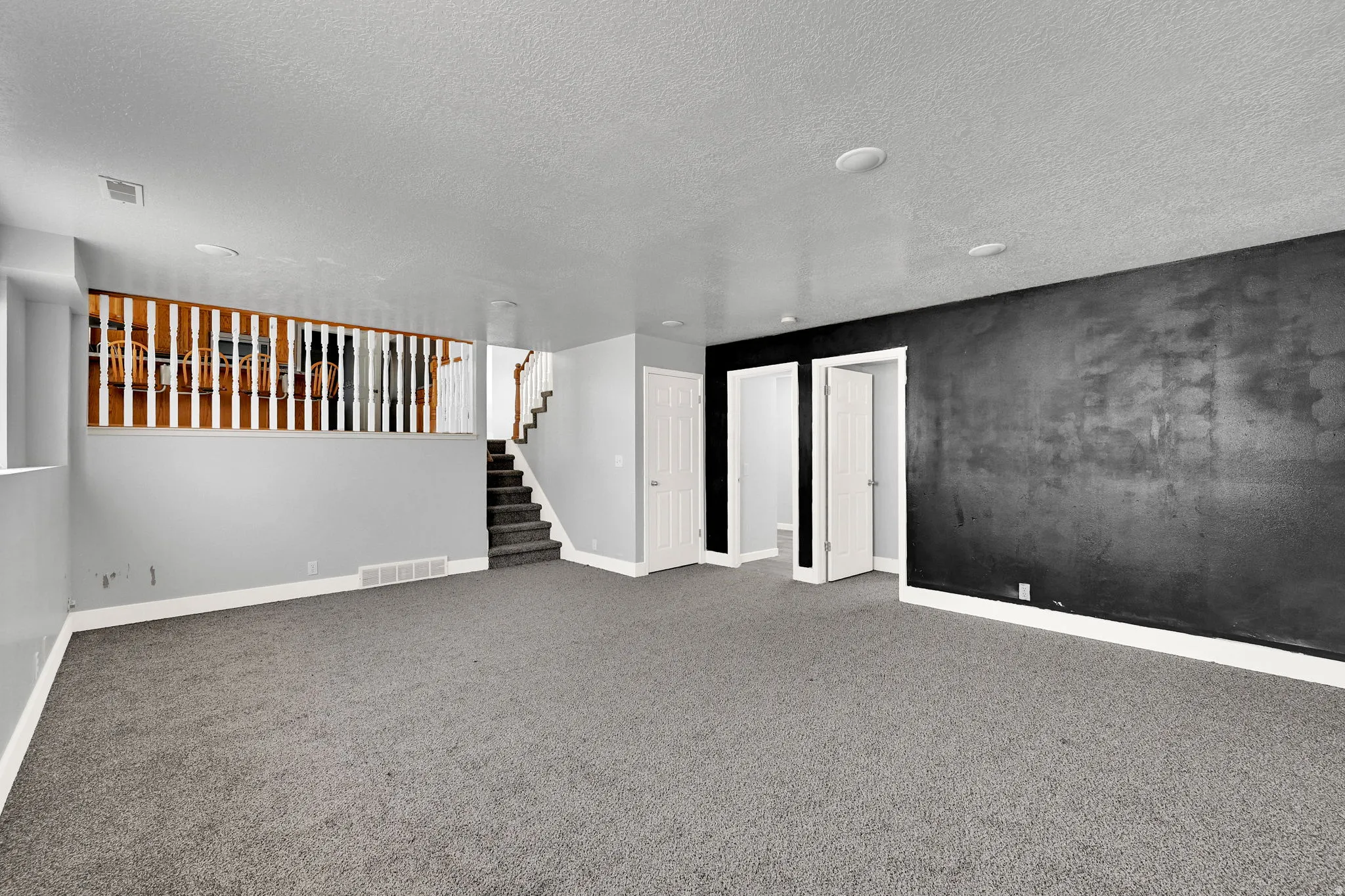 Spacious carpeted room featuring a dark accent wall, recessed lighting, and a staircase with a wood and white spindle railing