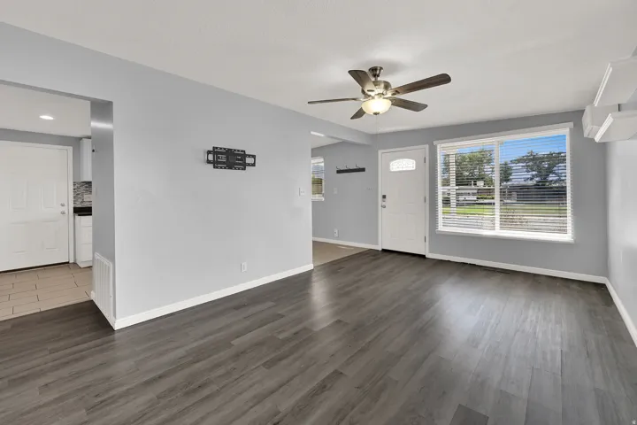 Open living space featuring wood-finish flooring, light gray walls, a ceiling fan with integrated lighting, and a white paneled entry door with a decorative glass insert