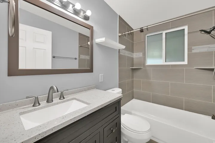 Bathroom featuring a vanity with a light-toned speckled countertop, brushed nickel fixtures, a framed mirror, a tiled shower/tub combination with integrated shelving, and a window for natural light