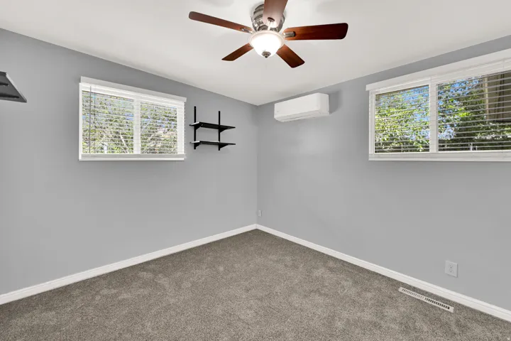 Room featuring two windows with blinds, a ceiling fan with integrated lighting, a wall-mounted air conditioning unit, and built-in shelving