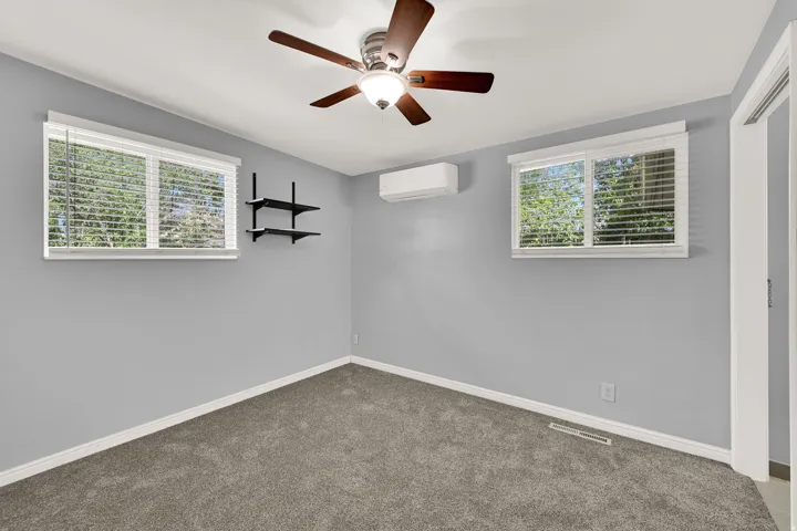 Carpeted room featuring a ceiling fan with integrated lighting, a wall-mounted mini-split HVAC unit, two windows with blinds, and white trim