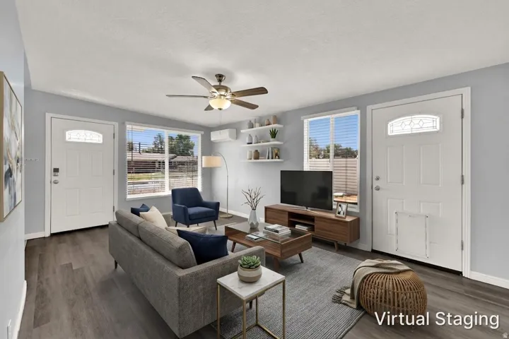 Open-concept living area featuring wood-finish flooring, a ceiling fan with integrated lighting, multiple windows with blinds, a wall-mounted mini-split system, and built-in floating shelves