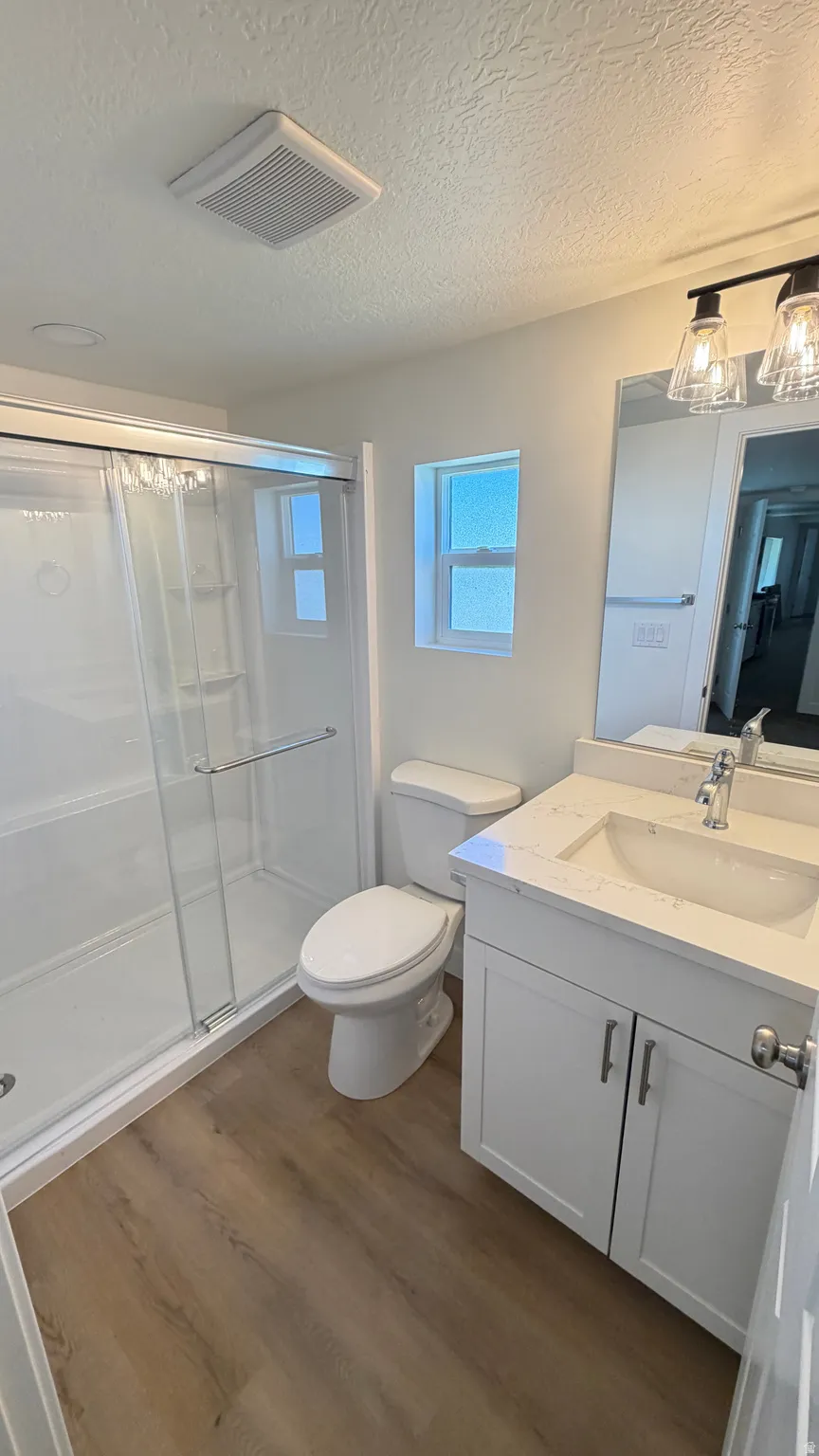 Bathroom featuring a white vanity with a light-toned countertop, a shower with a sliding glass door, wood-finish flooring, a toilet, and a window