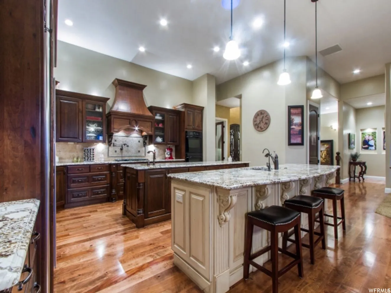 Spacious kitchen featuring two islands with stone countertops, a decorative wood range hood, rich wood cabinetry, and wood-finish flooring