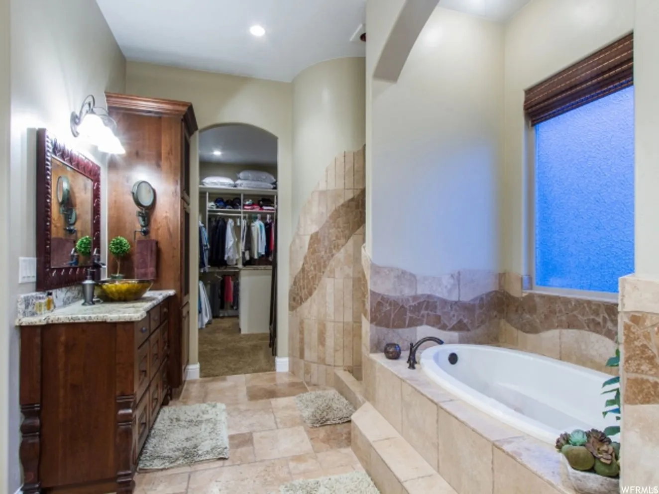 Spacious bathroom featuring a built-in tub, tiled surround, wood vanity with a stone countertop, and an arched doorway leading to a walk-in closet