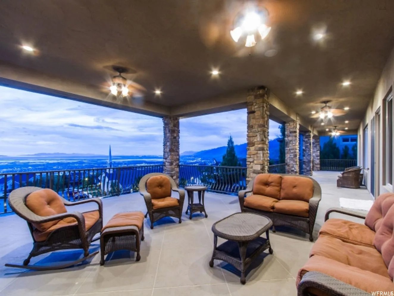 Expansive covered patio featuring multiple ceiling fans with integrated lighting, recessed ceiling lights, stone-clad support columns, and a railing overlooking a distant landscape