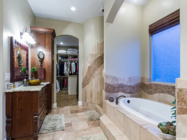 Spacious bathroom featuring a built-in tub, tiled surround, wood vanity with a stone countertop, and an arched doorway leading to a walk-in closet
