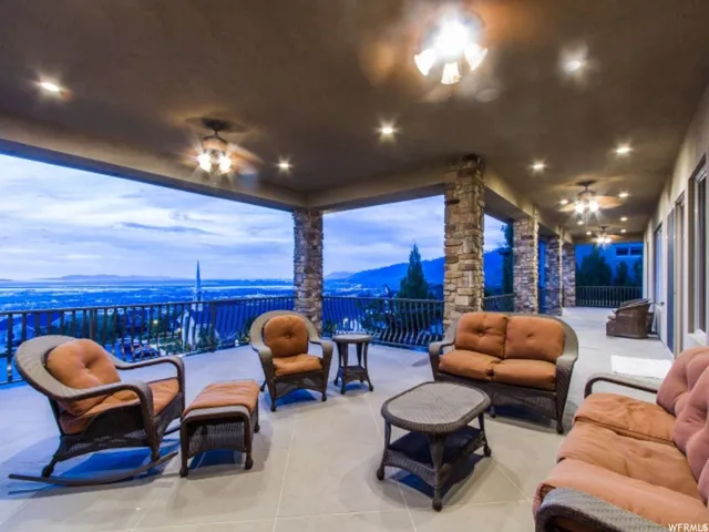 Expansive covered patio featuring multiple ceiling fans with integrated lighting, recessed ceiling lights, stone-clad support columns, and a railing overlooking a distant landscape