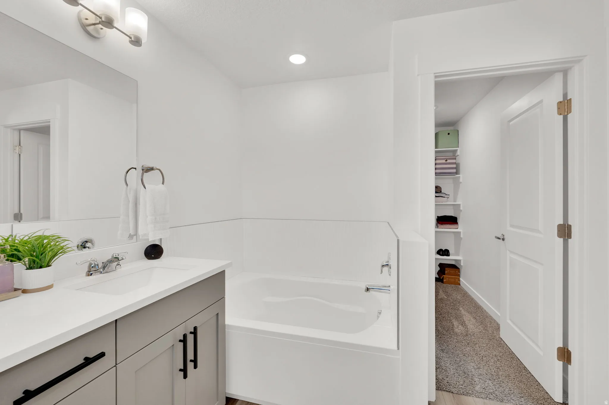 White bathroom featuring an integrated bathtub, a single vanity with a light gray cabinet and white countertop, a wall-mounted mirror, and an overhead light fixture