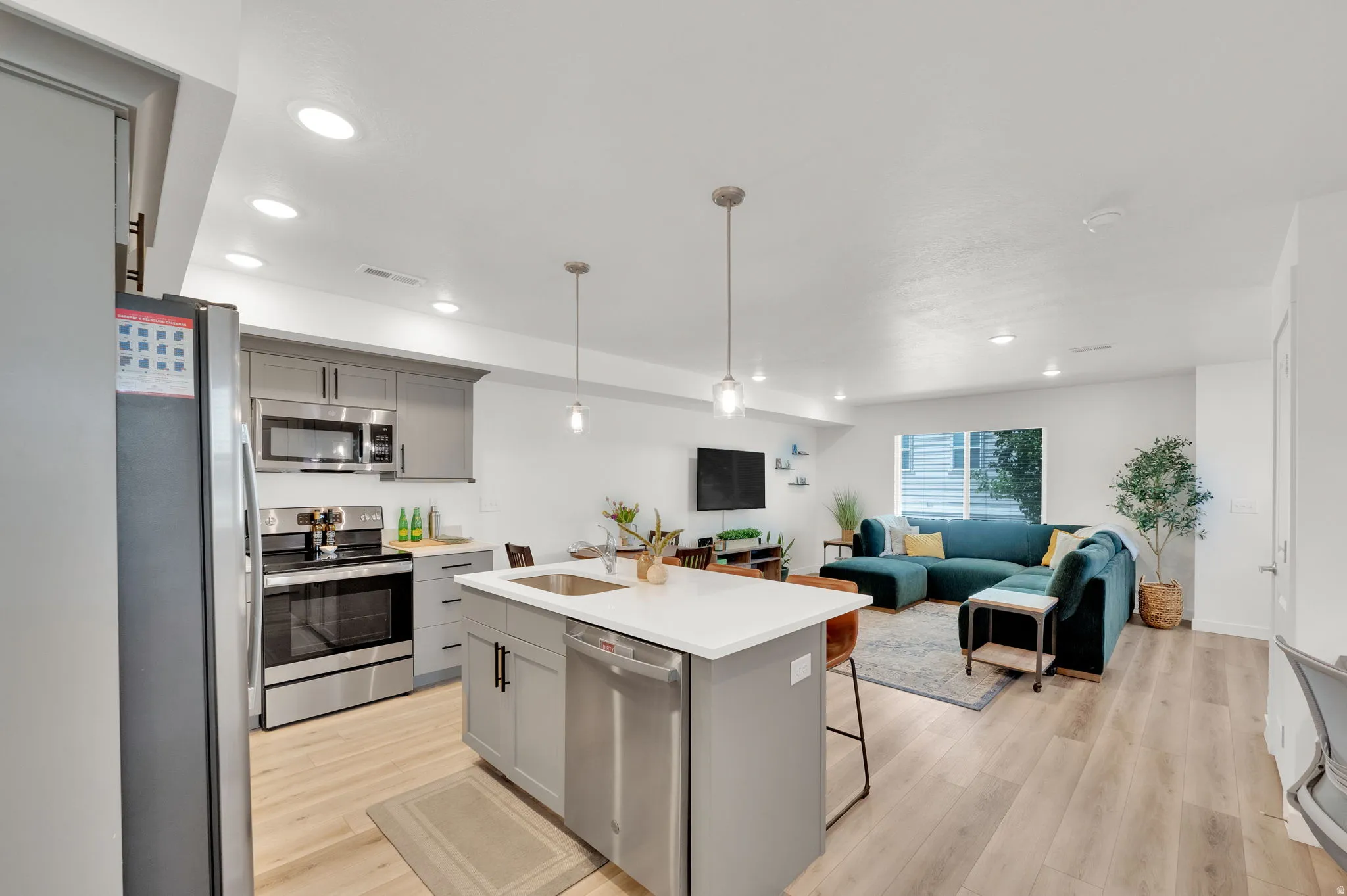 Open-concept living space with light wood-finish flooring, a kitchen island featuring a sink and pendant lighting, stainless steel appliances, grey cabinetry, and recessed lighting throughout