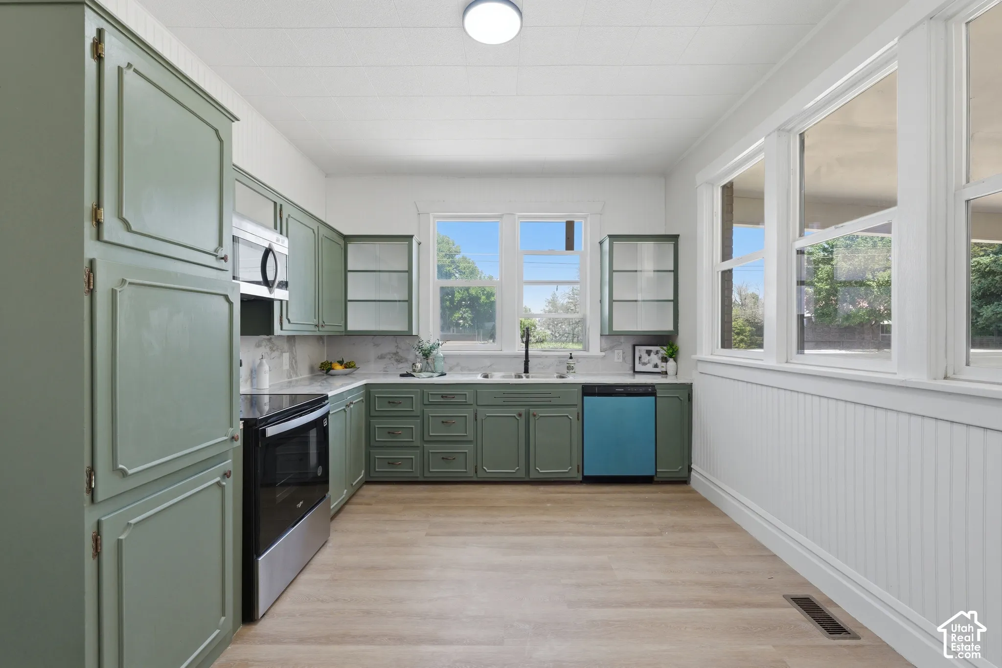 Kitchen featuring sage green cabinetry, stainless steel oven and microwave, light wood-finish flooring, white beadboard wall paneling, and multiple windows