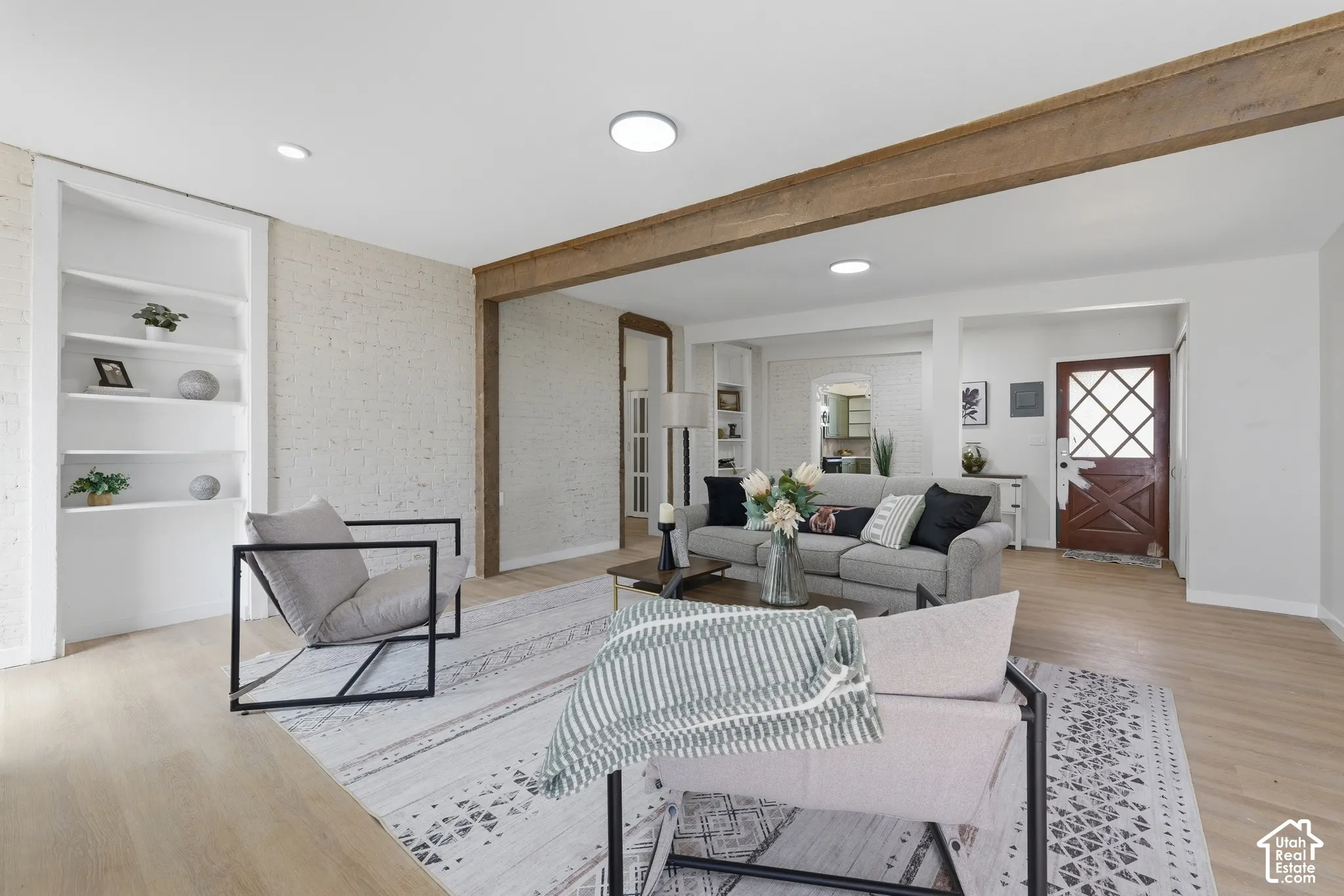 Spacious living area featuring wood-finish flooring, exposed ceiling beams, recessed lighting, a white brick accent wall, and built-in shelving