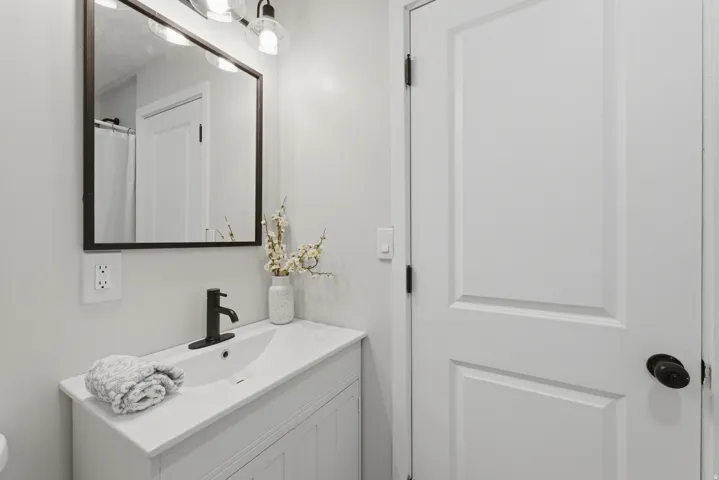 White vanity with an integrated sink and matte black faucet, complemented by a rectangular framed mirror and three-light vanity fixture