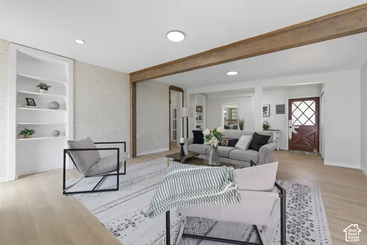 Spacious living area featuring wood-finish flooring, exposed ceiling beams, recessed lighting, a white brick accent wall, and built-in shelving