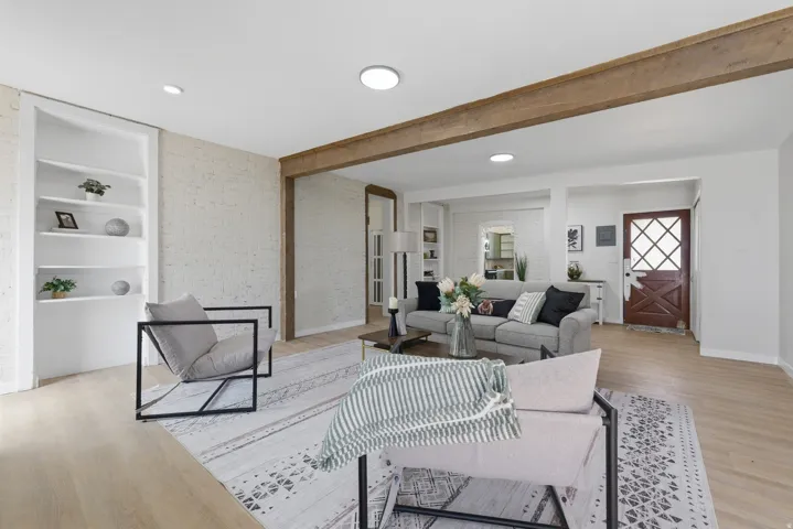 Spacious living area featuring wood-finish flooring, exposed ceiling beams, recessed lighting, a white brick accent wall, and built-in shelving