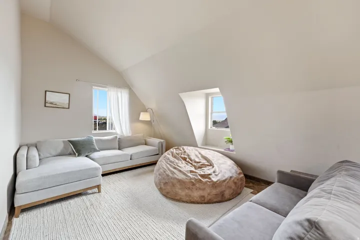 Attic living space featuring an angled ceiling, two windows, and a light-toned area rug over wood-finish flooring