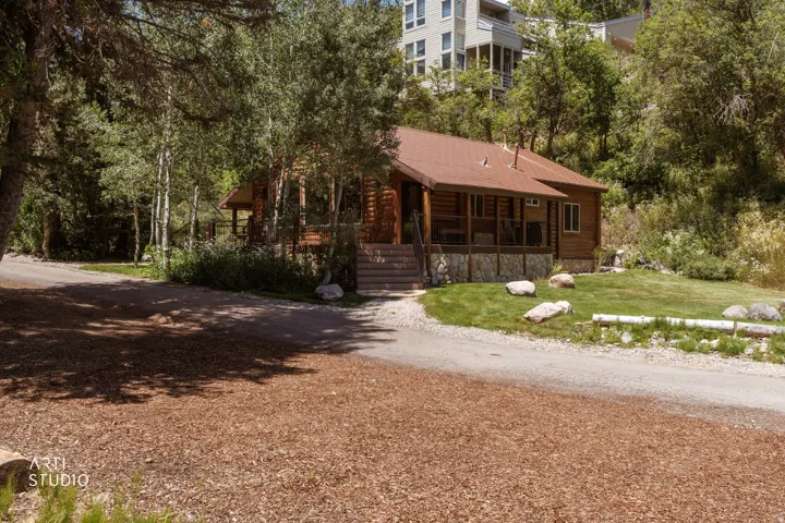 Rustic log cabin exterior with a brown aluminum metal roof, covered front porch, gravel driveway with space for 4 cars, and a verdant lawn surrounded by mature trees