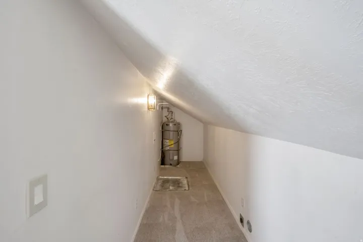 Carpeted storage area featuring an installed wall sconce, water heater, and a sloped ceiling