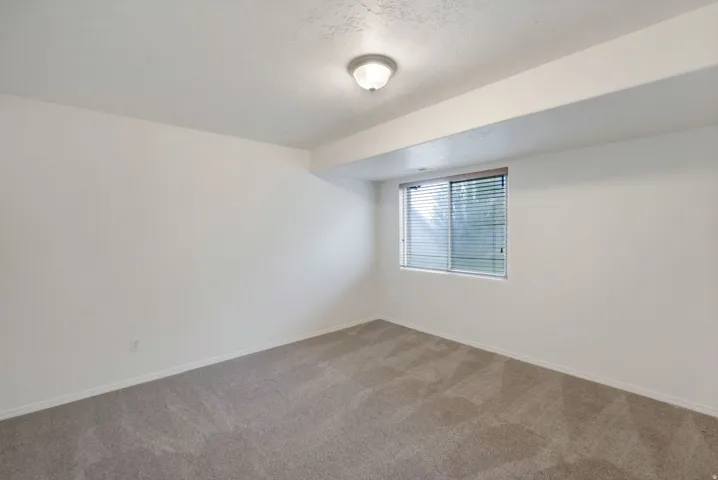 Spacious interior room featuring light-colored walls, neutral carpet flooring, and a single window with blinds