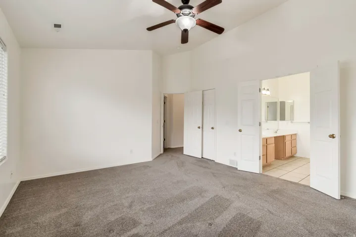 Spacious carpeted room featuring a vaulted ceiling with a ceiling fan, a window with horizontal blinds, and double doors opening to a bathroom with dual vanities