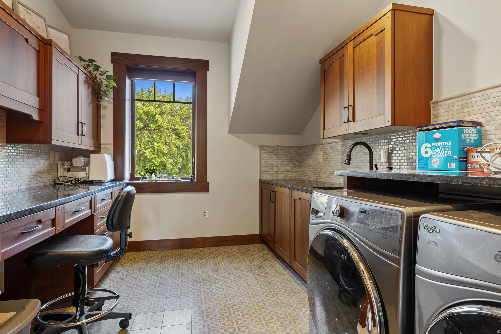 Laundry room featuring a built-in desk area, extensive wood cabinetry, dark countertops, a tile backsplash, and patterned floor tiling