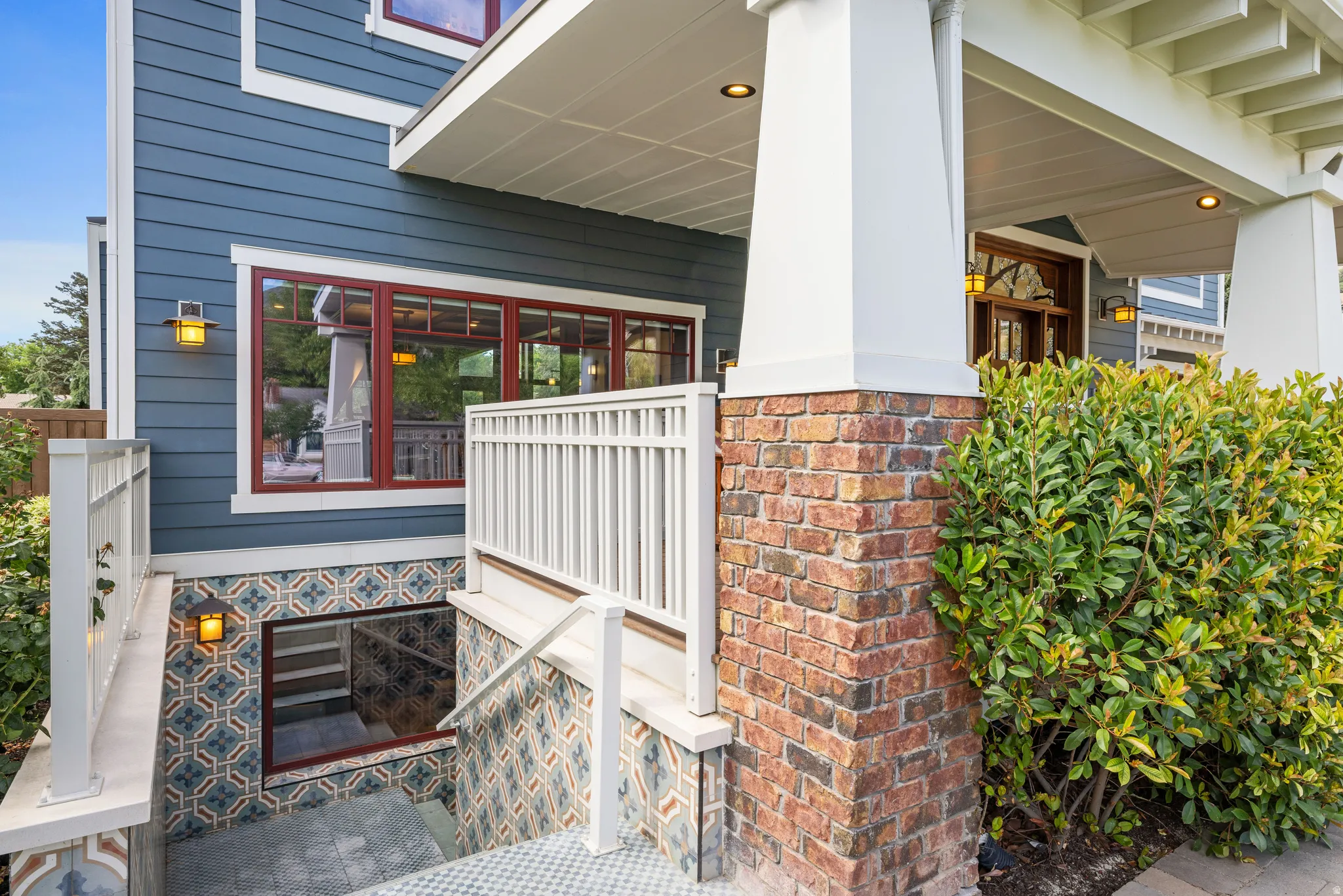 Exterior entry featuring blue horizontal siding, white architectural trim, brick column base, and a tiled lower entryway with recessed lighting