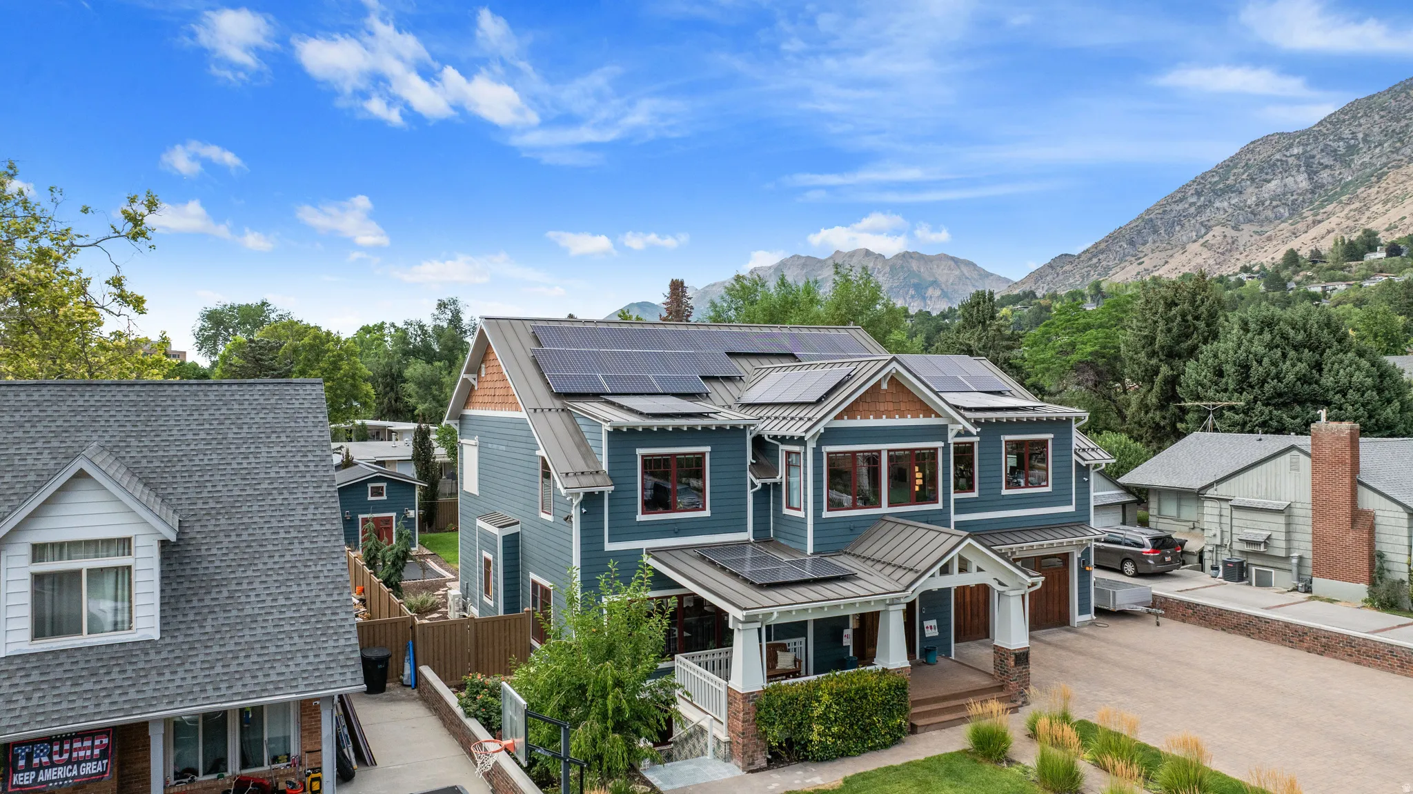 Multi-story residence featuring blue siding, a covered front porch with stone pillars, a two-car garage with wood-finish doors, multiple solar panel arrays, and a mountain backdrop