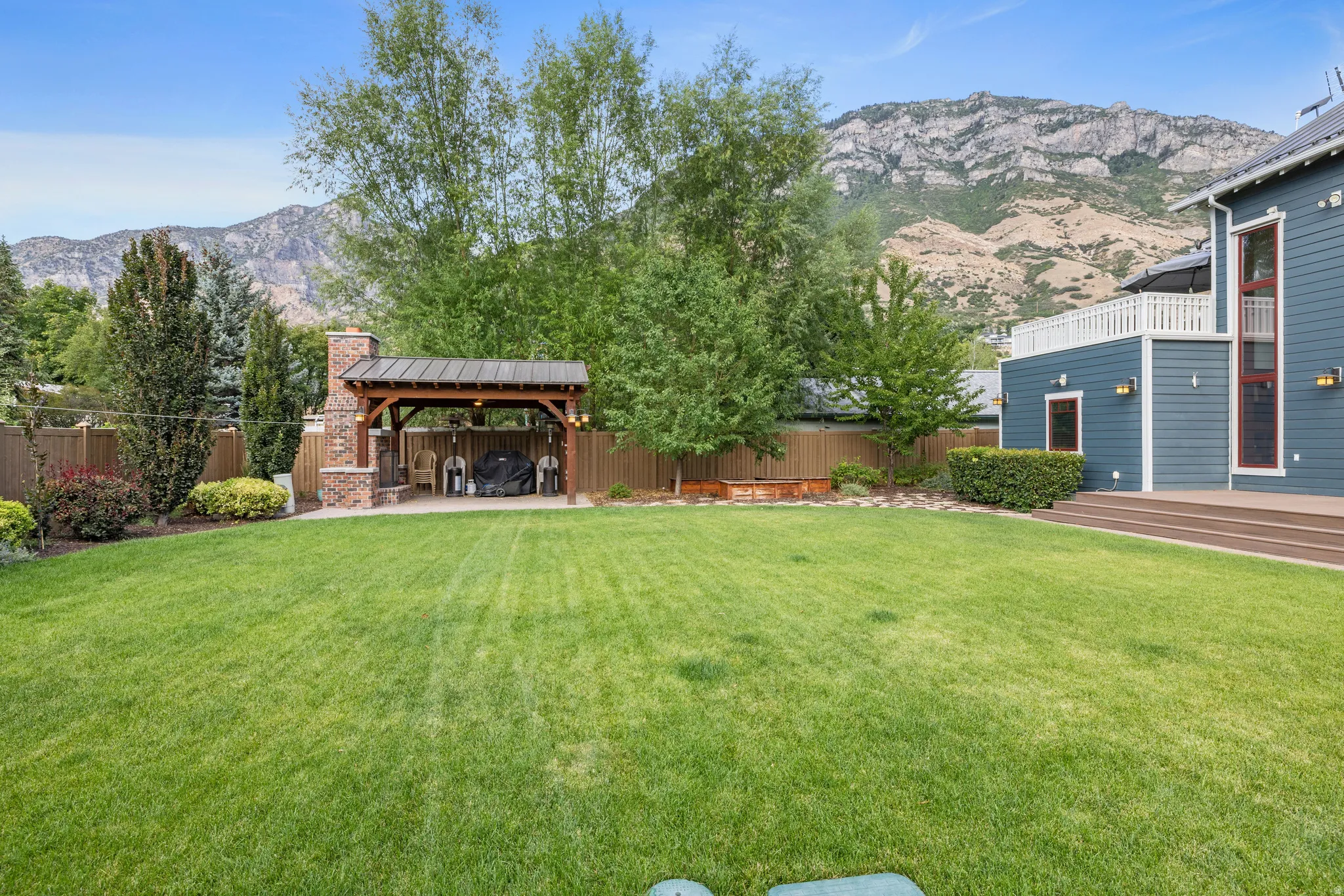 Expansive lawn featuring a stone-clad outdoor fireplace, a covered patio structure, and mature trees